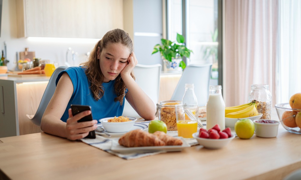 Una adolescente mirando el móvil pensativa frente al desayuno